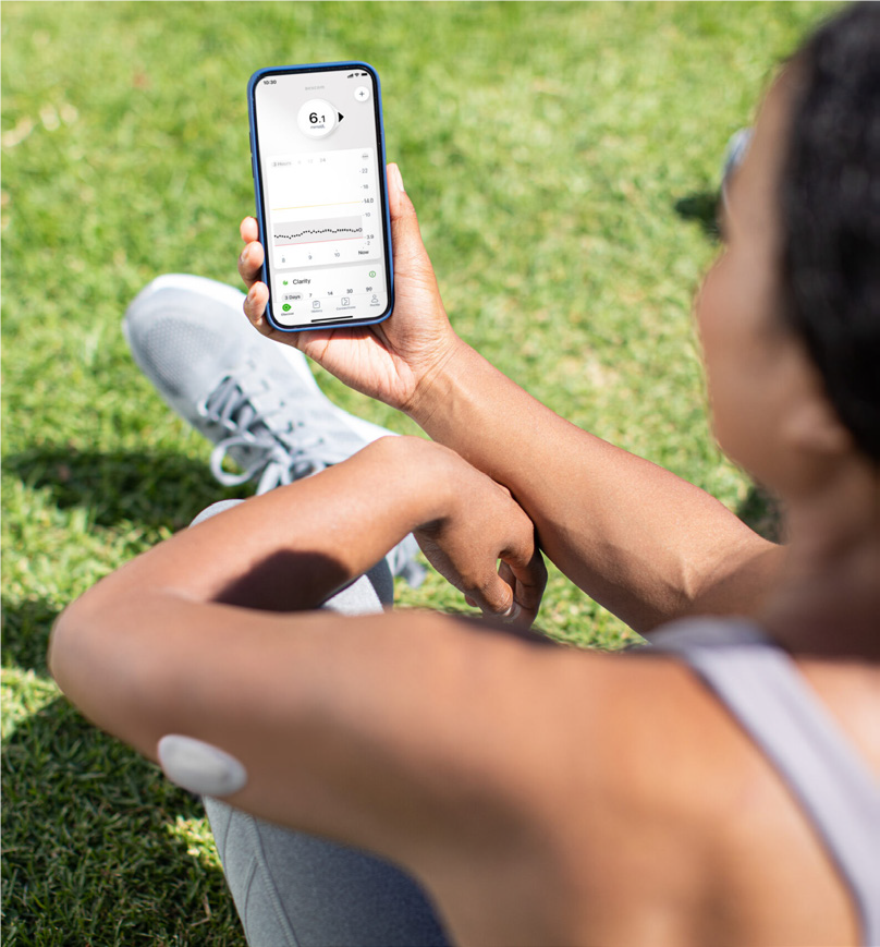 We zien een vrouw die tijdens het sporten even buiten op het gras is gaan zitten in de zon. ze kijkt haar glocuse na via de app die verbonden is met haar meter in haar arm.
